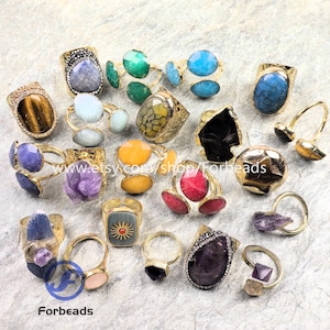 May include: A selection of gold-coloured rings with diverse gemstone settings. The rings showcase a range of colours, including blue, green, yellow, purple, and red. Some rings have a textured appearance, while others are smooth. The rings are displayed on a light grey surface.