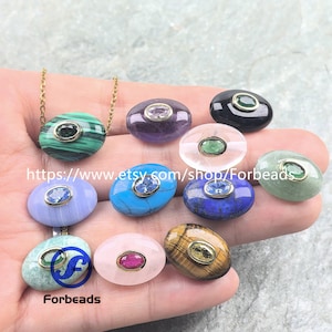 Puede incluir: Una colección de colgantes de piedras preciosas de forma ovalada en varios colores, incluyendo verde, morado, azul y rosa, cada uno con un bisel dorado. Un colgante está unido a una cadena dorada. Los colgantes se muestran en una mano.