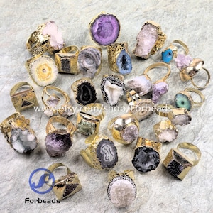 May include: A selection of gold-coloured rings with diverse natural stone settings. The stones exhibit a spectrum of colours, such as purple, white, blue, and green. The rings have unique, irregular shapes and textures, with some featuring a geode-like appearance.