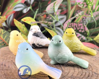 May include: Five decorative bird ornaments in various colours, including yellow, blue, green, and white, displayed on a wooden surface. The birds have a glossy finish and are set against a backdrop of green and pink foliage. The text "Forbeads" is visible.