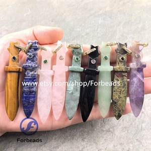 Puede incluir: Una colección de nueve colgantes con forma de daga tallados en varias piedras preciosas, incluyendo ojo de tigre, lapislázuli, cuarzo rosa y amatista. Cada colgante presenta un engaste dorado con pequeños cristales y una piedra incrustada.