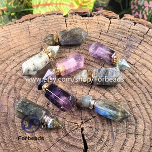 May include: A collection of gemstone essential oil bottles in shades of purple, pink, and grey. Each bottle features a gold or silver cap and a pointed crystal top. The bottles are displayed on a rustic wooden surface. The Etsy shop name "Forbeads" is visible.