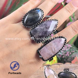 May include: Close-up of three gemstone rings. Each ring features a different colored stone: a dark gray, a purple, and a light pink. The stones are set in a dark metal with a beaded border. The rings are on a hand.