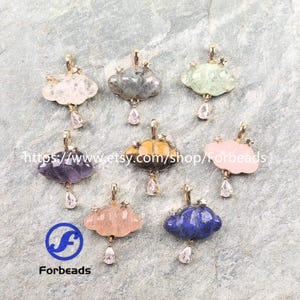 Pode incluir: Uma coleção de pingentes em forma de nuvem em várias cores, incluindo rosa, roxo e azul. Cada pingente tem uma argola dourada, pequenos detalhes de estrelas e um cristal em forma de gota. Os pingentes são exibidos em uma superfície cinza texturizada.