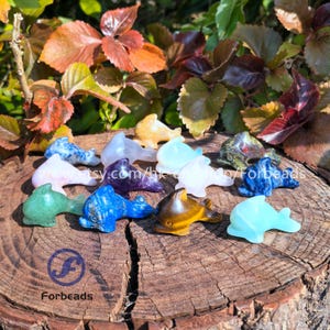 May include: A collection of 12 hand-carved dolphin figurines made from various types of polished gemstones. The dolphins are in a variety of colours, including green, blue, purple, pink, brown, and white. The dolphins are arranged in a circle on a wooden surface.