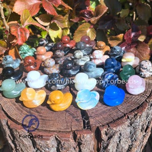 May include: A collection of 25 hand-carved gemstone duck figurines in various colors and styles. The ducks are made from natural stones like amethyst, agate, jasper, and more. They are arranged on a wooden surface.