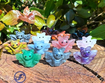 Puede incluir: Una colección de 12 murciélagos de cristal tallados a mano en varios colores, incluyendo verde, morado, rosa, gris y blanco. Los murciélagos están dispuestos sobre una superficie de madera.