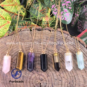 May include: A collection of seven crystal pendant necklaces with gold-colored chains. The crystals are various colors, including pink, yellow, purple, black, clear, and green. The necklaces are displayed on a wooden surface with green foliage in the background. The Etsy shop name "Forbeads" is visible.