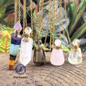 May include: A collection of perfume bottle necklaces with gold chains. The bottles are various colors, including clear, smoky gray, and pink, and are topped with gold-colored stoppers. The necklaces are displayed on a wooden surface with green foliage in the background. The text "www.etsy.com/shop/Forbeads" is visible.