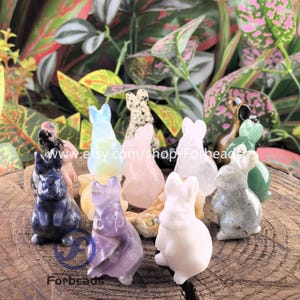 May include: A collection of small, carved bunny figurines in various colors and materials, including blue, pink, and white. The bunnies are displayed on a wooden surface with green foliage in the background. The Etsy shop name "Forbeads" is visible.