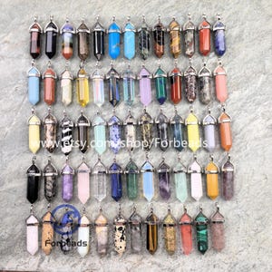 Pode incluir: Uma coleção de pingentes de pedras preciosas em várias cores, incluindo preto, azul, verde e laranja. Cada pingente tem uma tampa prateada e uma forma pontiaguda semelhante a um cristal. Os pingentes estão dispostos em uma superfície cinza.