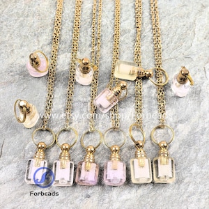 Puede incluir: Una colección de pequeños colgantes de botellas de perfume de vidrio transparente con detalles metálicos dorados y cadenas. Las botellas, rectangulares y redondas, contienen líquido rosa o amarillo. Los colgantes están dispuestos sobre una superficie texturizada.