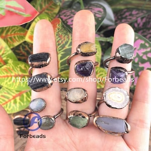 Op de afbeelding: Een verzameling edelsteenringen in verschillende kleuren, waaronder zwart, paars, geel en groen. De ringen hebben een koperkleurige band en worden op vingers getoond. De Etsy shopnaam "Forbeads" is zichtbaar.