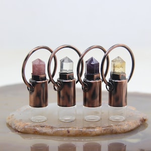 May include: Four small glass bottles with crystal toppers in shades of pink, clear, purple, and yellow. Each bottle has a copper-colored metal base and a circular handle. The bottles are arranged on a light-colored, patterned surface.