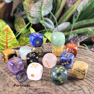 May include: A collection of gemstone dice in various colors, including blue, green, red, and purple, with gold-colored dots. The dice are displayed on a wooden surface with green foliage in the background. The text "www.etsy.com/shop/Forbeads" is visible.