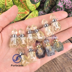Puede incluir: Una colección de pequeños colgantes con forma de frasco de perfume facetado, hechos de piedra translúcida. Cada frasco cuenta con una tapa metálica con una pequeña anilla para colgar. Las piedras varían en color, incluyendo tonos amarillo pálido, gris humo y morado.