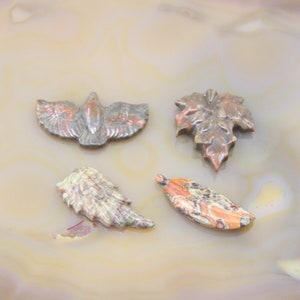 May include: Four carved stone pendants in the shape of leaves and a bird. The pendants are made of a variety of colors, including brown, green, and orange.