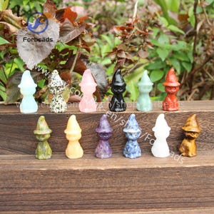 May include: A collection of 14 small, carved crystal figurines in various colors, each depicting a witch wearing a pointed hat. The figurines are arranged in a row on a wooden surface.