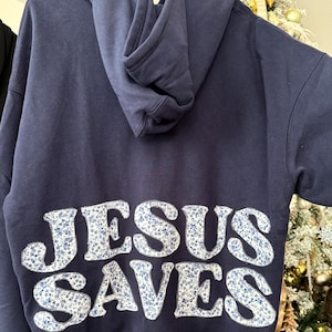 May include: Navy blue hoodie with a hood. The words "JESUS SAVES" are appliqued on the back in white fabric with a blue floral pattern. The hoodie is made of a soft material.
