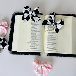 May include: A black and white checkered bow, a pink and white checkered bow, and a pink and white gingham bow are used as bookmarks in a large, open book with black fringe.