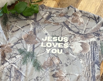 May include: Camouflage-patterned t-shirt with the text "JESUS LOVES YOU" in white block letters. The shirt features a woodland camouflage design with brown, green, and gray tones. The brand name "HOLLISTER" is visible on the collar.