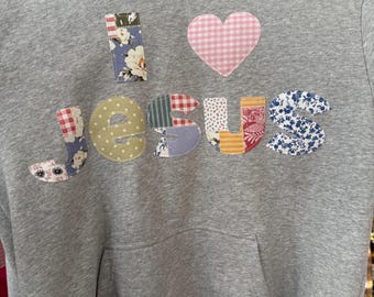May include: A gray hoodie with a front pocket and a hood. The words "I LOVE JESUS" are appliqued on the front in colorful, patterned fabrics, including floral and gingham designs. A pink gingham heart is above the word "JESUS".