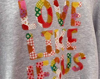 May include: A gray hoodie with the words "LOVE LIKE JESUS" appliquéd across the front. The letters are made of colorful patchwork fabric with floral, polka dot, and checkered patterns. The hoodie has a hood and a kangaroo pocket.