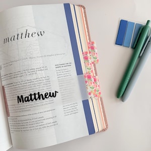 May include: Open Bible with the word "Matthew" prominently displayed. The pages are marked with floral patterned tabs and blue and light blue sticky notes. Two pens, one green and one gray, are also visible.