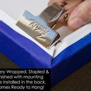 May include: A canvas art piece with blue and white edges being secured with a silver metal clamp. The image includes the text: "Gallery Wrapped, Stapled & Finished with mounting wire installed in the back. Comes Ready to Hang!"