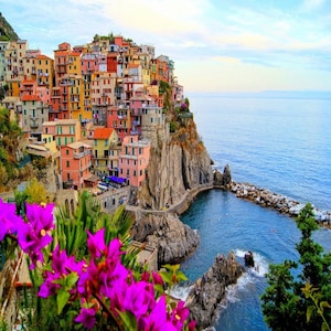 May include: A picturesque coastal village with brightly coloured buildings clinging to a cliff face. The buildings are painted in shades of pink, yellow, and orange. Purple flowers bloom in the foreground, with the sea a deep blue.