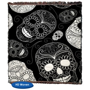 May include: Black and white throw blanket with a repeating pattern of sugar skulls with intricate floral designs. The blanket has a brown fringe border and the words "HD Woven" are printed on a blue tag.