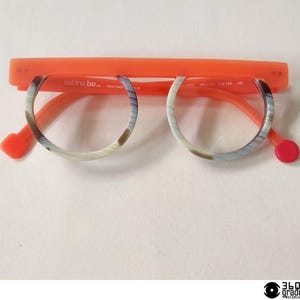 Puede incluir: Gafas con montura naranja y lentes circulares con un patrón veteado en tonos grises y marrones. Las patillas y el puente nasal son naranjas, con un acento rojo en las puntas de las patillas. El texto "sabine be.." es visible.