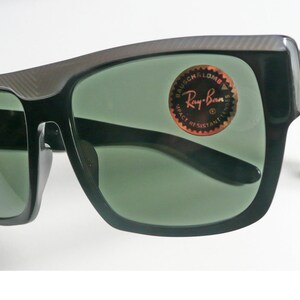 May include: Black Ray-Ban sunglasses with a carbon fiber frame and green lenses. The sunglasses have a sticker on the right lens that says "Ray-Ban" and "Impact Resistant".
