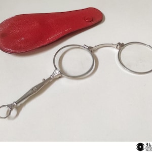 Op de afbeelding: Zilverkleurige, ronde bril met een handvat en een rode leren etui. De bril heeft heldere glazen en een decoratief handvat. De rode etui is ovaal van vorm en heeft een knoopsluiting. De bril is waarschijnlijk vintage of antiek.