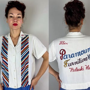 Puede incluir: Camisa blanca de manga corta con detalles de rayas coloridas y el texto "Paramount Furniture Co" bordado en la espalda. La camisa tiene cuello y cierre de botones. El texto "Pulaski Hwy" también está bordado en la espalda.