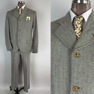 Puede incluir: Un traje de espiga gris claro con chaqueta y pantalón a juego. La chaqueta tiene solapas con muescas, tres botones y un pañuelo de bolsillo. El traje se exhibe en un maniquí. La corbata tiene un patrón único.