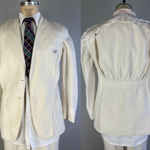 May include: An off-white blazer featuring a two-button front closure, a chest pocket with a small label, and long sleeves. The back displays a gathered waist detail with a horizontal seam. A white collared shirt and a dark tie with a grid pattern.