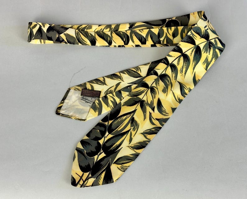 May include: A yellow silk tie with a black and green leaf pattern. The tie has a brown label that reads "Debron's, Hand Made Neckwear, New York".