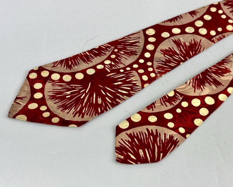 May include: A red tie with a repeating pattern of beige circles and white dots. The tie has a shiny finish and a classic shape. The design features a radial pattern with a vintage aesthetic.