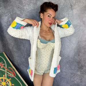 May include: A white textured cardigan featuring long sleeves with yellow, blue, and pink floral accents at the cuffs and lower pockets. The garment is worn over a light blue and white polka dot bodysuit with a structured bodice and thin straps.