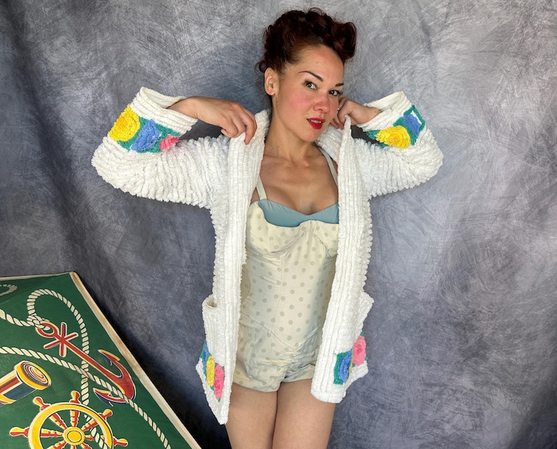 May include: A white textured cardigan featuring long sleeves with yellow, blue, and pink floral accents at the cuffs and lower pockets. The garment is worn over a light blue and white polka dot bodysuit with a structured bodice and thin straps.