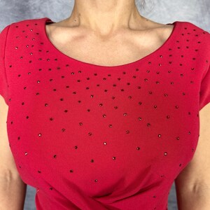 May include: A bright red short-sleeved top featuring a wide scoop neckline and a scattered pattern of small, dark, reflective embellishments across the chest and shoulder areas, creating a subtle textured appearance on the fabric surface.