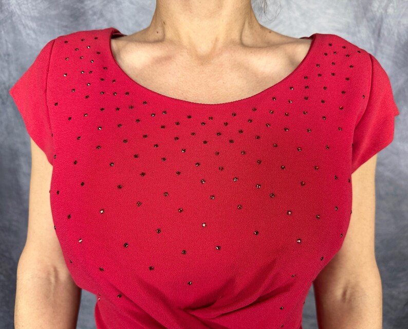 May include: A bright red short-sleeved top featuring a wide scoop neckline and a scattered pattern of small, dark, reflective embellishments across the chest and shoulder areas, creating a subtle textured appearance on the fabric surface.