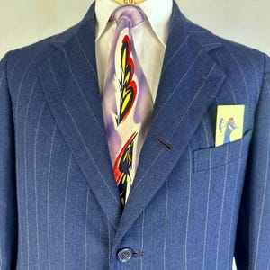 Puede incluir: Un blazer azul de rayas finas sobre una camisa blanca de cuello y una corbata con un patr&oacute;n abstracto de plumas en morado, amarillo y rojo. Un peque&ntilde;o pa&ntilde;uelo de bolsillo amarillo con una ilustraci&oacute;n de un pavo real azul en el bolsillo.