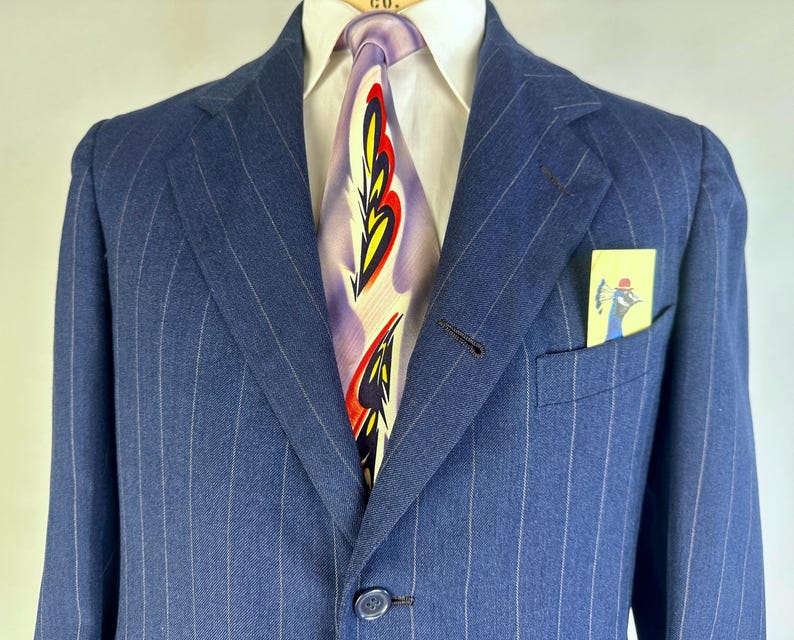 Puede incluir: Un blazer azul de rayas finas sobre una camisa blanca de cuello y una corbata con un patr&oacute;n abstracto de plumas en morado, amarillo y rojo. Un peque&ntilde;o pa&ntilde;uelo de bolsillo amarillo con una ilustraci&oacute;n de un pavo real azul en el bolsillo.