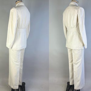 May include: A two-piece off-white suit comprising a long-sleeved jacket with a cinched waist and matching straight-leg trousers. The jacket features a high collar, structured shoulders, and buttoned cuffs, while the trousers have a clean hem.