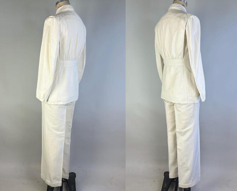 May include: A two-piece off-white suit comprising a long-sleeved jacket with a cinched waist and matching straight-leg trousers. The jacket features a high collar, structured shoulders, and buttoned cuffs, while the trousers have a clean hem.