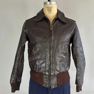 May include: A brown leather bomber jacket with a zip fastening and a ribbed knit collar, cuffs, and waistband. The jacket has a label that reads "Wolf Model Form Co. N.Y."