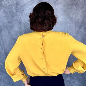 May include: A mustard yellow blouse with long sleeves and button details down the back. The sleeves have a gathered cuff detail. The blouse is paired with a dark blue skirt or pants. The model's back is facing the camera.