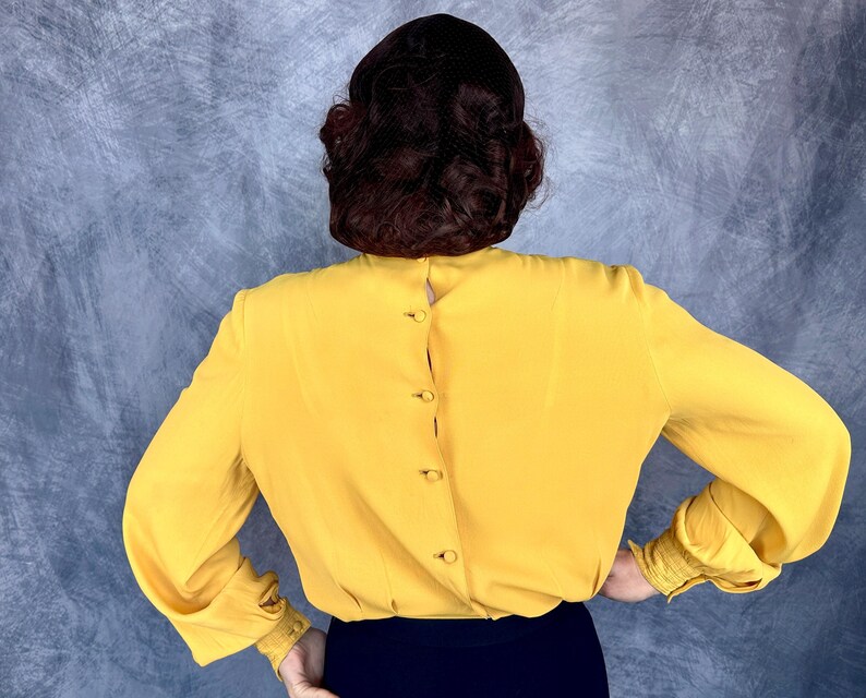 May include: A mustard yellow blouse with long sleeves and button details down the back. The sleeves have a gathered cuff detail. The blouse is paired with a dark blue skirt or pants. The model's back is facing the camera.